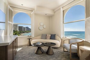 A modern living room with a sofa, two coffee tables, a chair, and large windows with a view of the ocean and buildings.