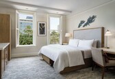 A hotel room with a large bed, two bedside tables with lamps, a chair, a window with shutters, and a wall-mounted artwork.