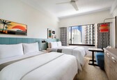 A hotel room with two beds, a ceiling fan, a window with a city view, and a painting on the wall.