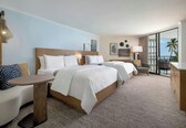 A hotel room with two beds, a sofa, a small table, and a balcony with a view of palm trees.