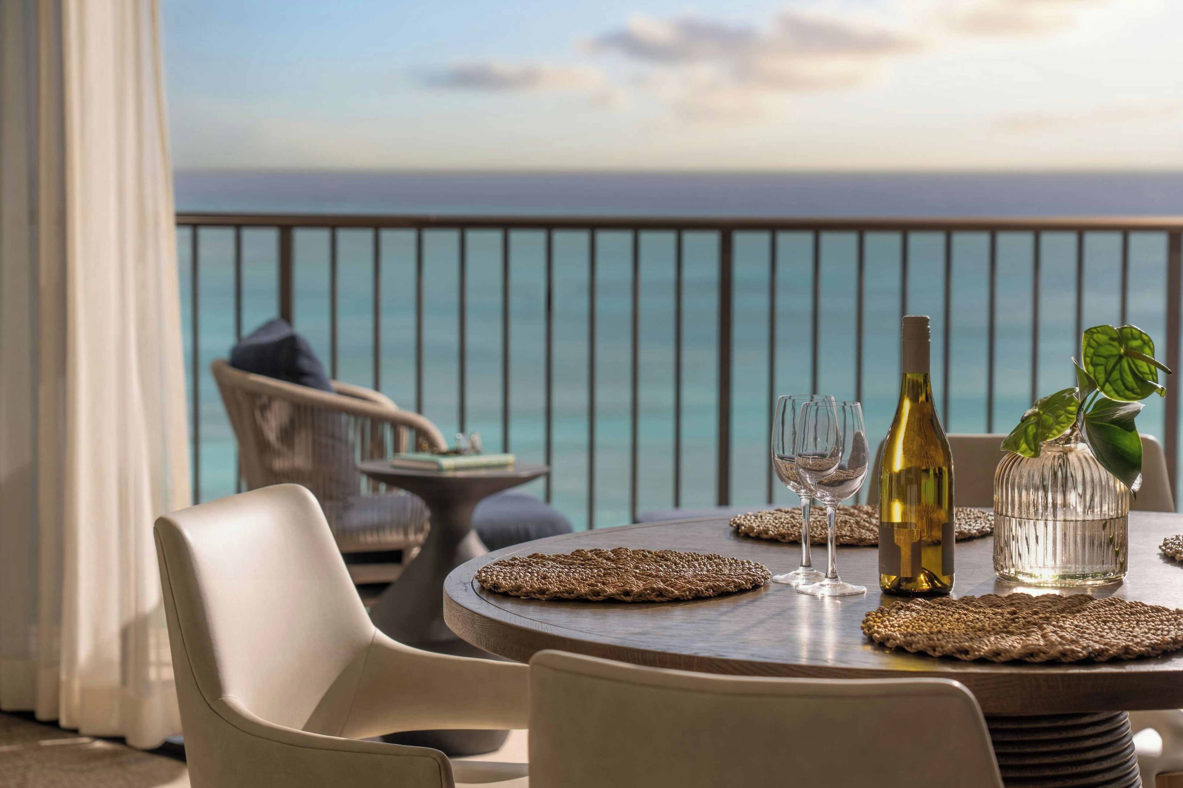 A round table with a bottle, glasses, and a plant on a balcony overlooking the sea.