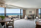 A spacious hotel room with a balcony, a round dining table, comfortable seating, and a large window offering a view of the sea.