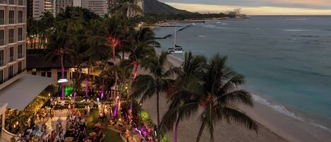 A rooftop bar with a view of the beach and ocean, surrounded by palm trees and hotel buildings.
