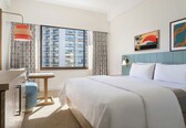 A hotel room with a large bed, a desk with a chair, a window with a view of a building, and artwork on the wall.
