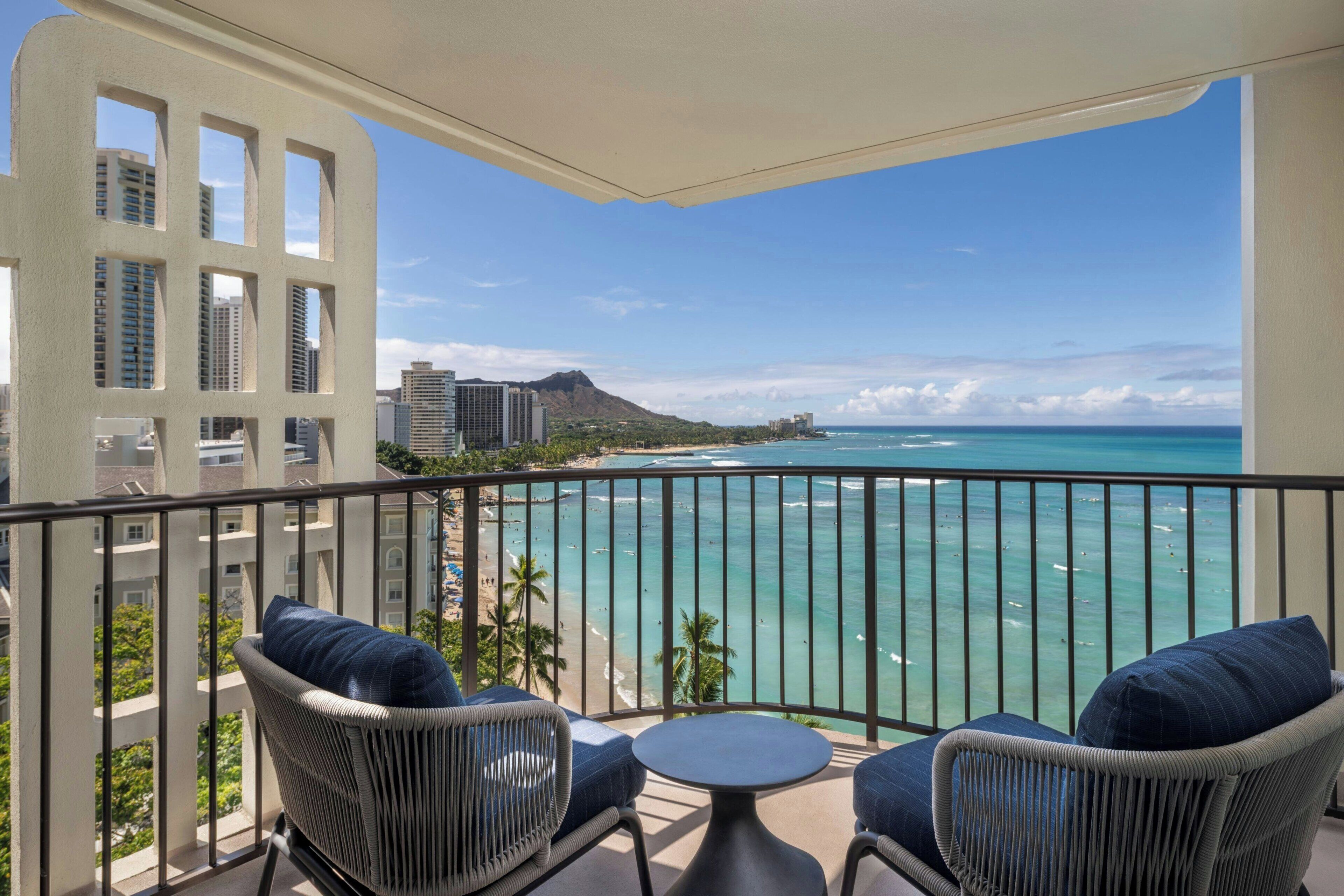 A balcony with wicker chairs and a small table, overlooking a beach with clear turquoise water and a mountain in the distance.