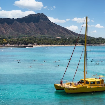 A yellow boat with "NA HOKU III" on its side, floating on clear turquoise water with people swimming and surfing in the background.