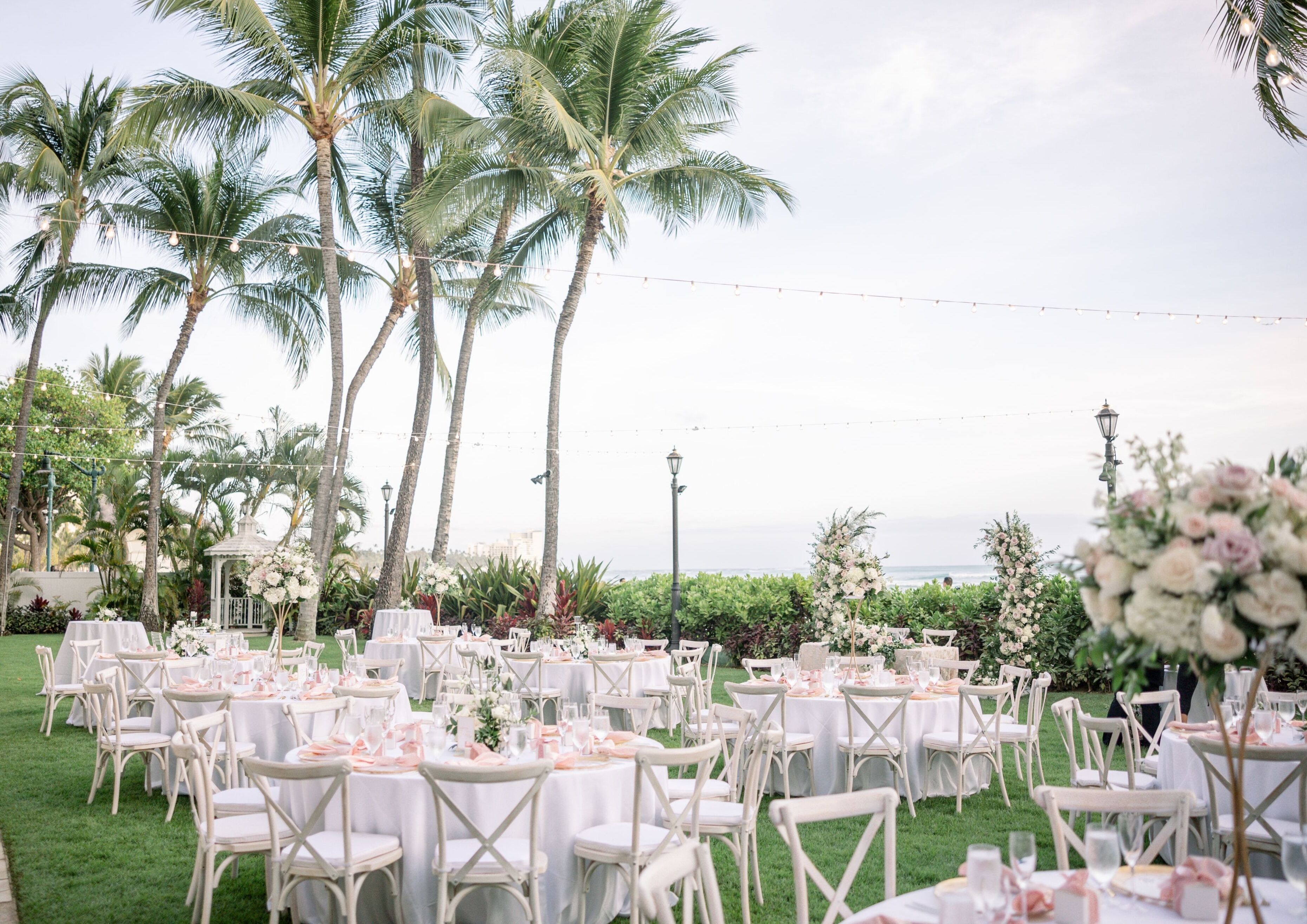 An outdoor wedding venue with round tables, white chairs, and string lights.