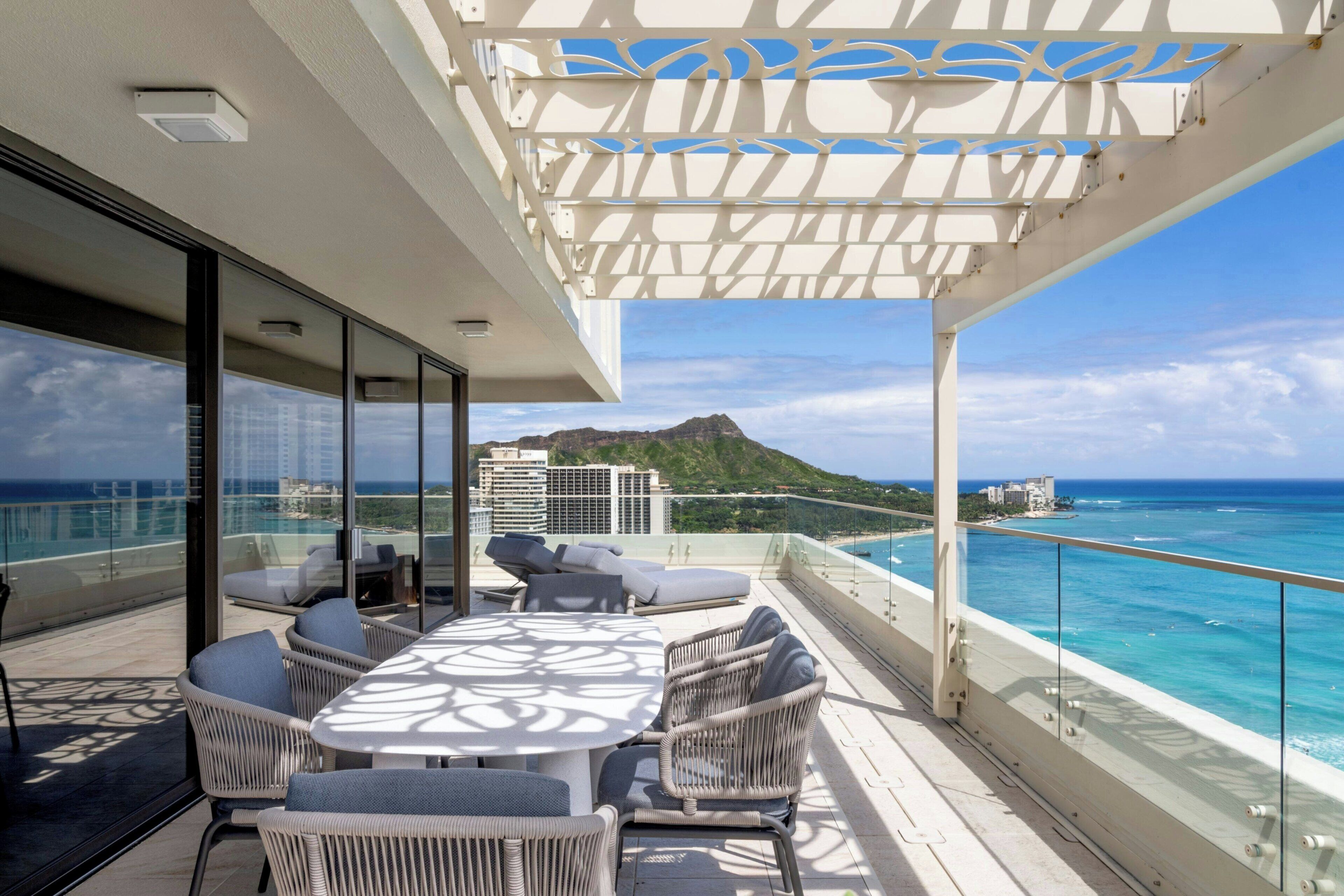 A balcony with a table and chairs, overlooking a coastal view with buildings and a mountain in the distance.