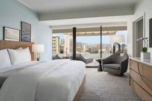 A hotel room with a bed, a chair, a desk, a lamp, and a view of the cityscape.