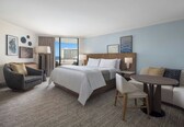 A hotel room with a large bed, a sofa, a round table, and a chair. There are two lamps, a coffee table, and a view of the city.