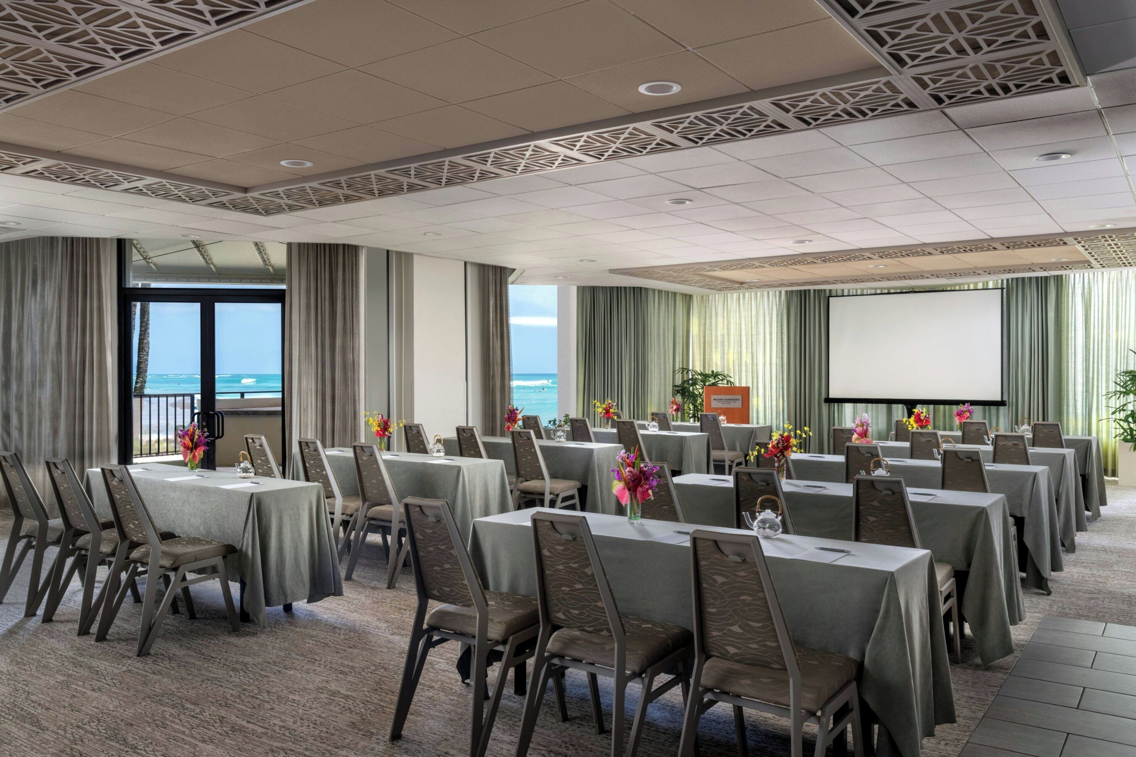 A conference room with tables and chairs, a projector screen, and a view of the ocean.