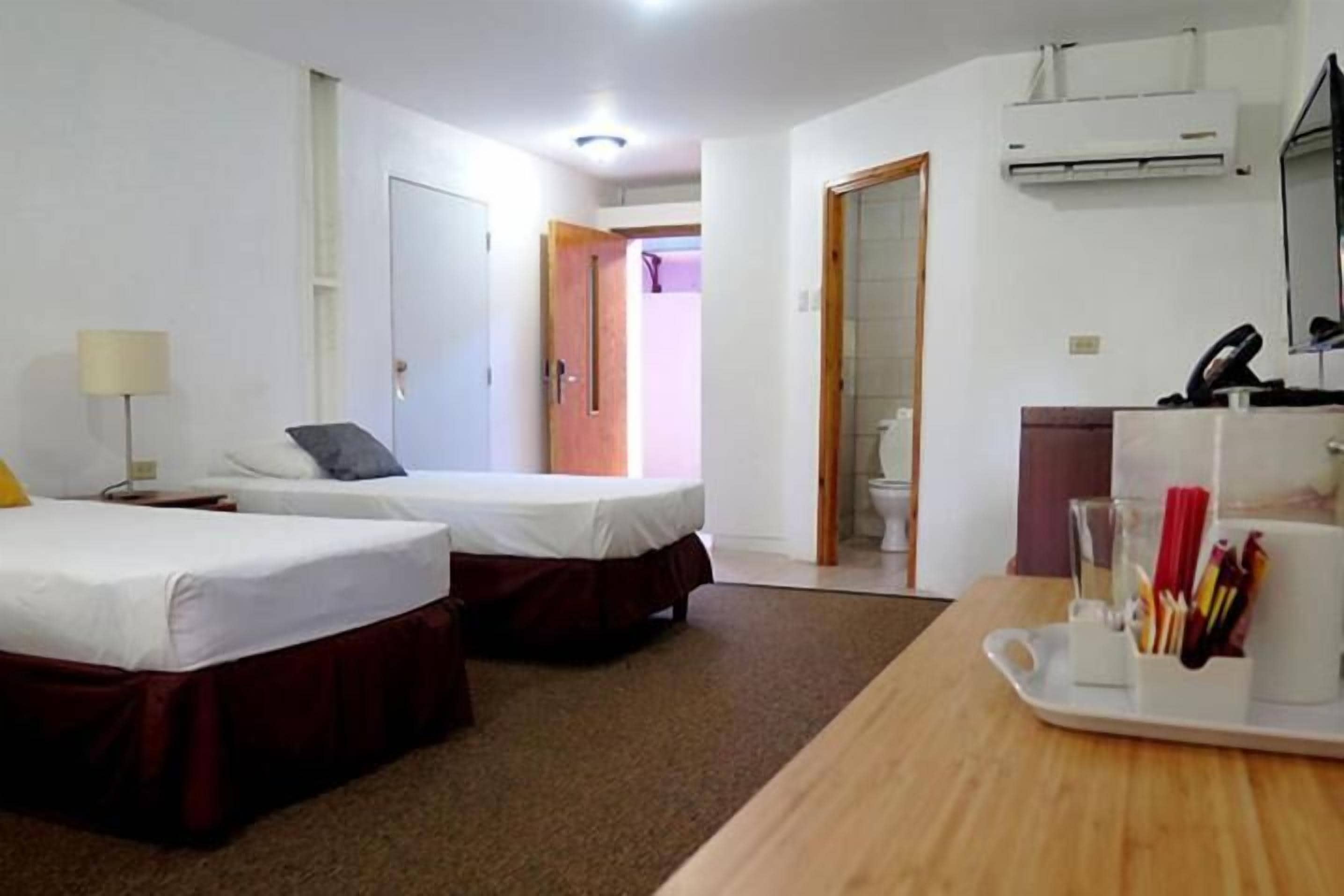 A hotel room with two beds, a desk with a tray of pens, a lamp, and an air conditioning unit.