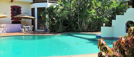 A pool area with a clear blue pool, surrounded by lush greenery and a two-story building with balconies.