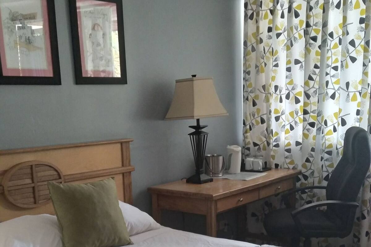 A hotel room with a large bed, a wooden headboard, a desk with a lamp, a chair, and a window with patterned curtains.