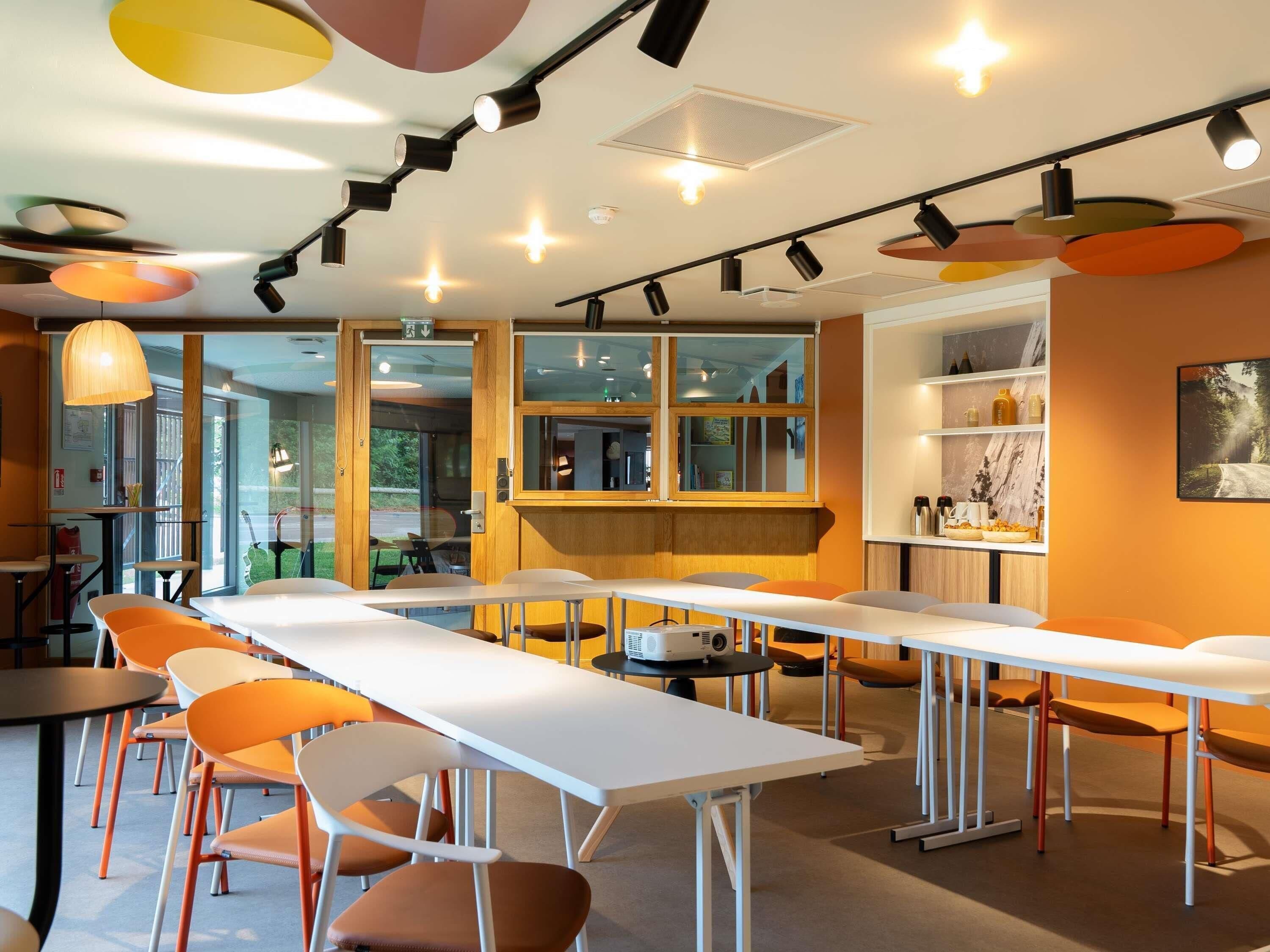 A modern dining area with long tables, orange chairs, and pendant lighting.