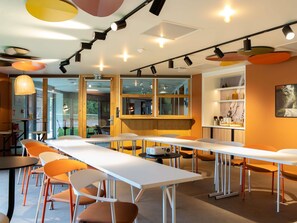 Un área de comedor moderna con mesas largas, sillas anaranjadas e iluminación colgante.