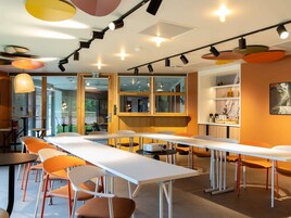 Un área de comedor moderna con mesas largas, sillas anaranjadas e iluminación colgante.