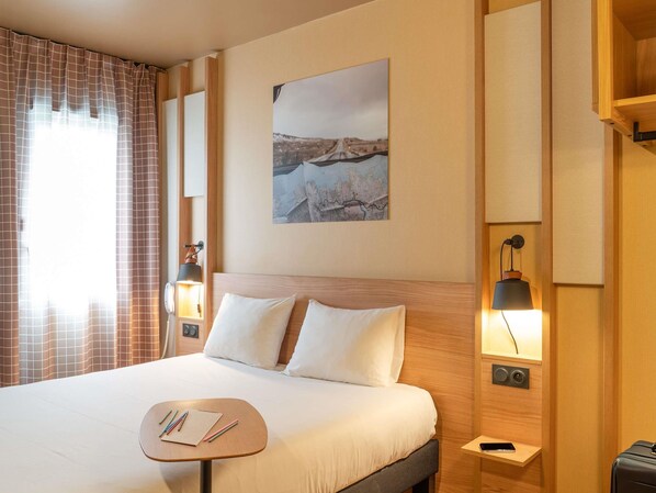 A hotel room with a bed, bedside lamps, a small table with stationery, a painting on the wall, and a window with curtains. - Ibis Styles Rouen Parc Expo Zenith (Saint-Etienne-du-Rouvray)