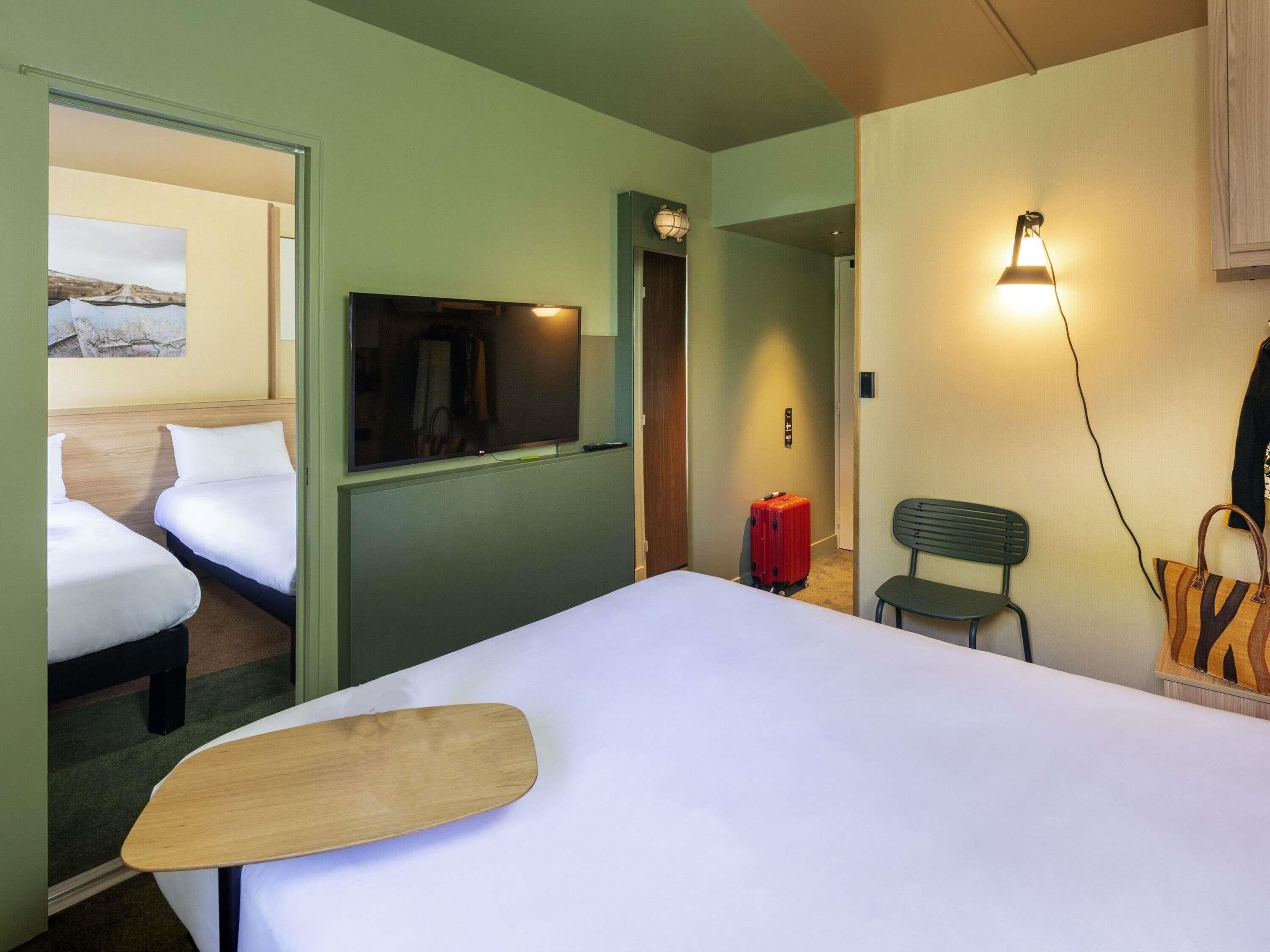 Une chambre d’hôtel avec un lit, une télévision, une valise, un banc et une lampe fixée au mur.