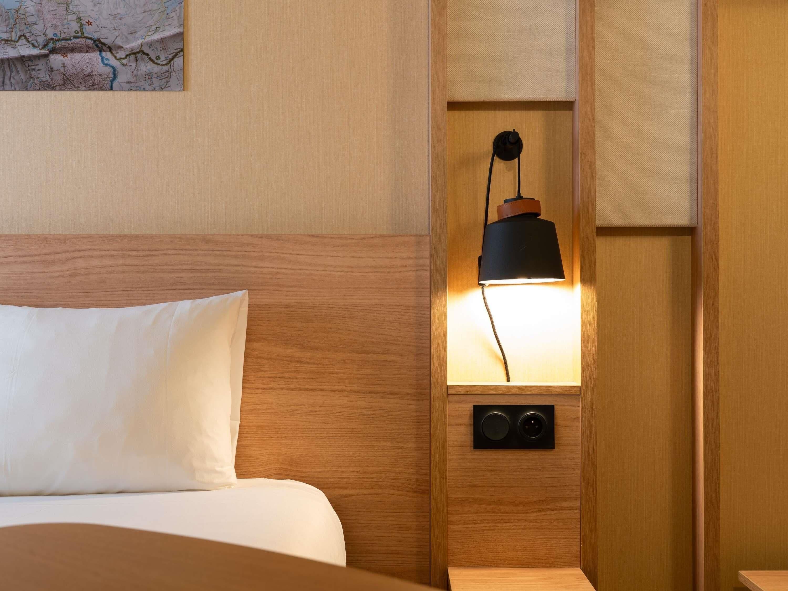 A hotel room with a wooden headboard, a bed with white bedding, a wall-mounted lamp, and a small speaker.