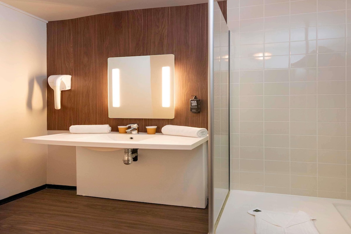 Une salle de bain moderne avec un mur en bois, un lavabo blanc, deux gobelets en papier, une serviette et une cabine de douche en verre.