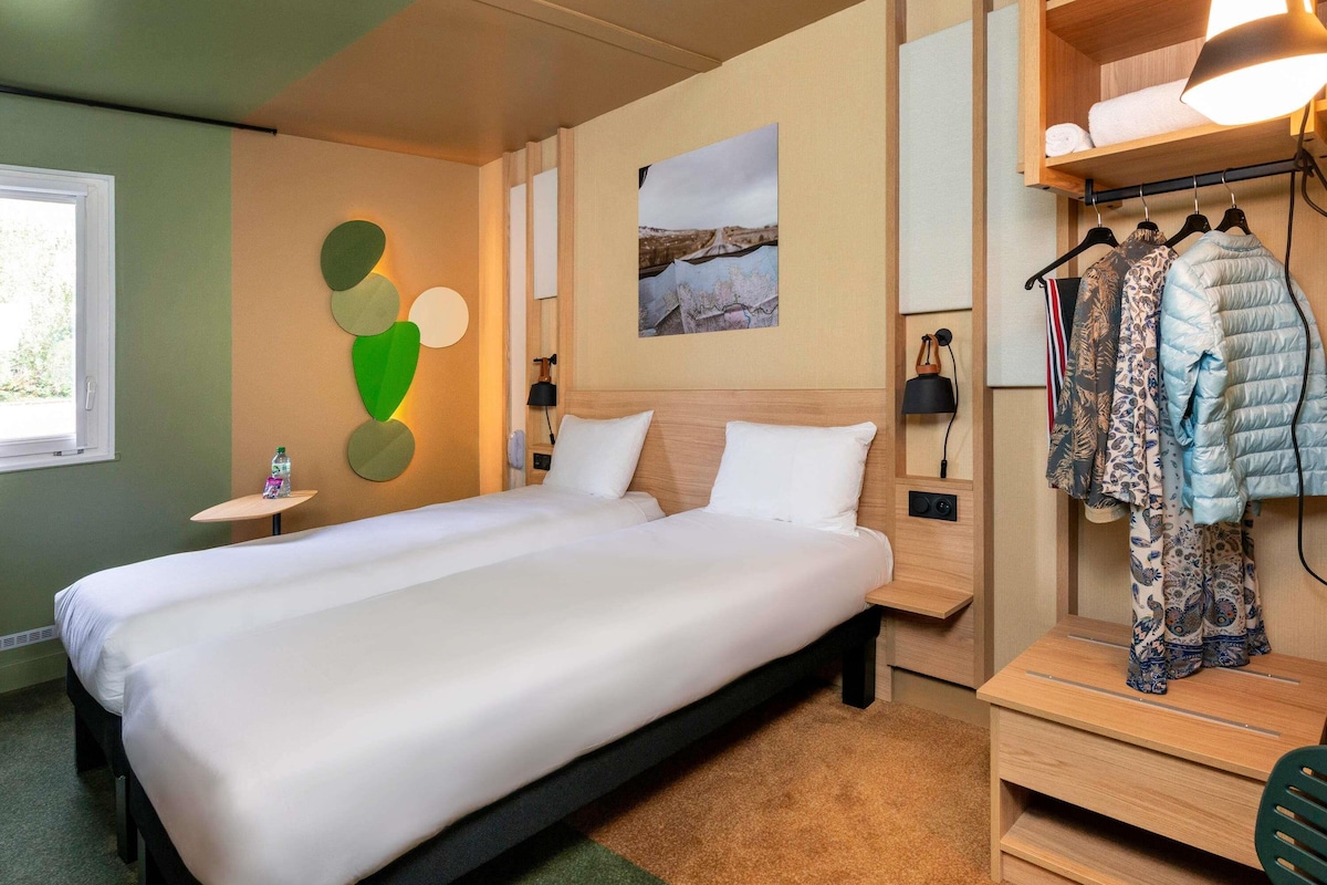 A hotel room with a large bed, a small bedside table, a wall-mounted lamp, a clothing rack, and a painting on the wall.