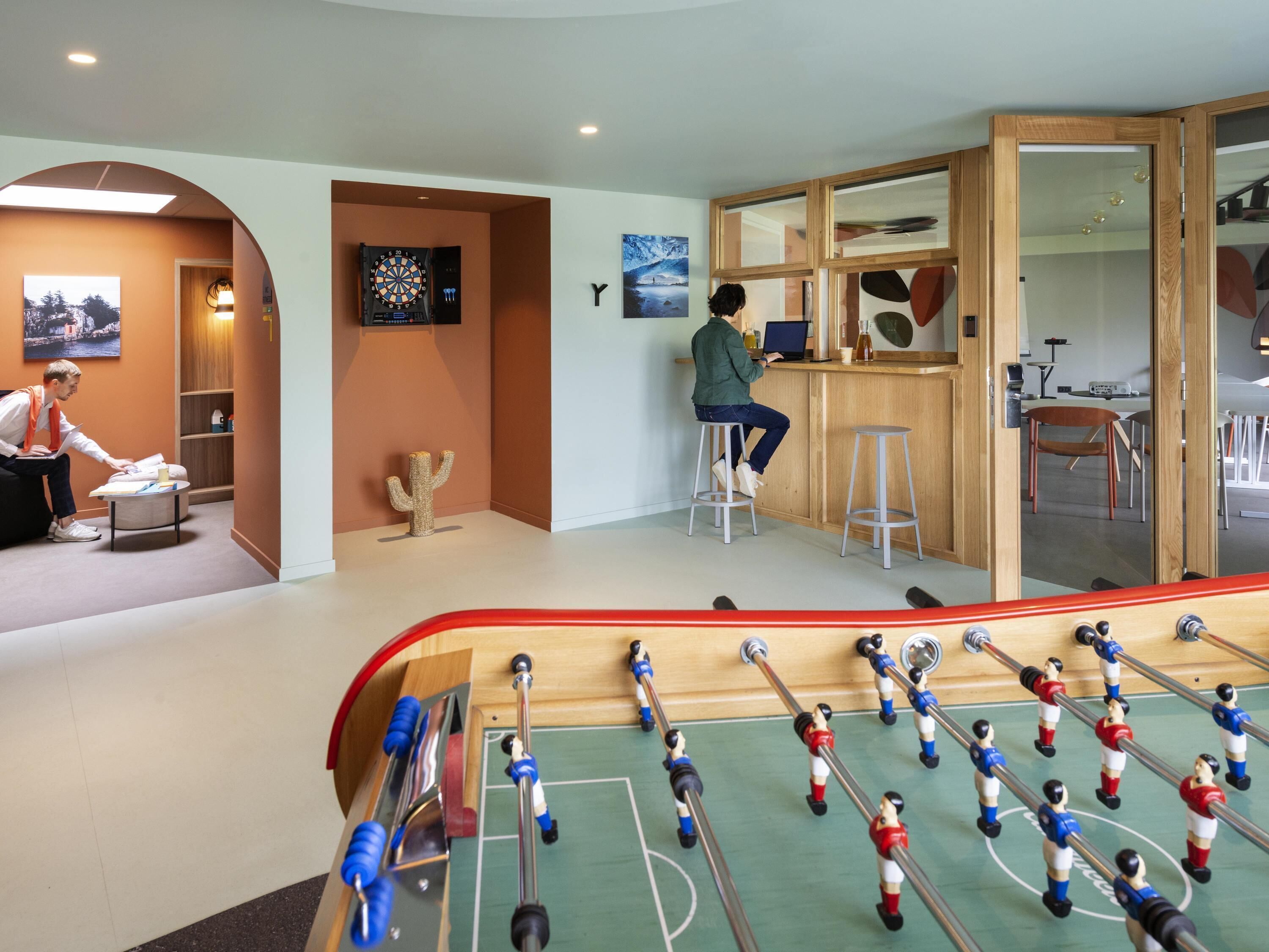 A room with a foosball table, a dartboard, and a person working at a desk.