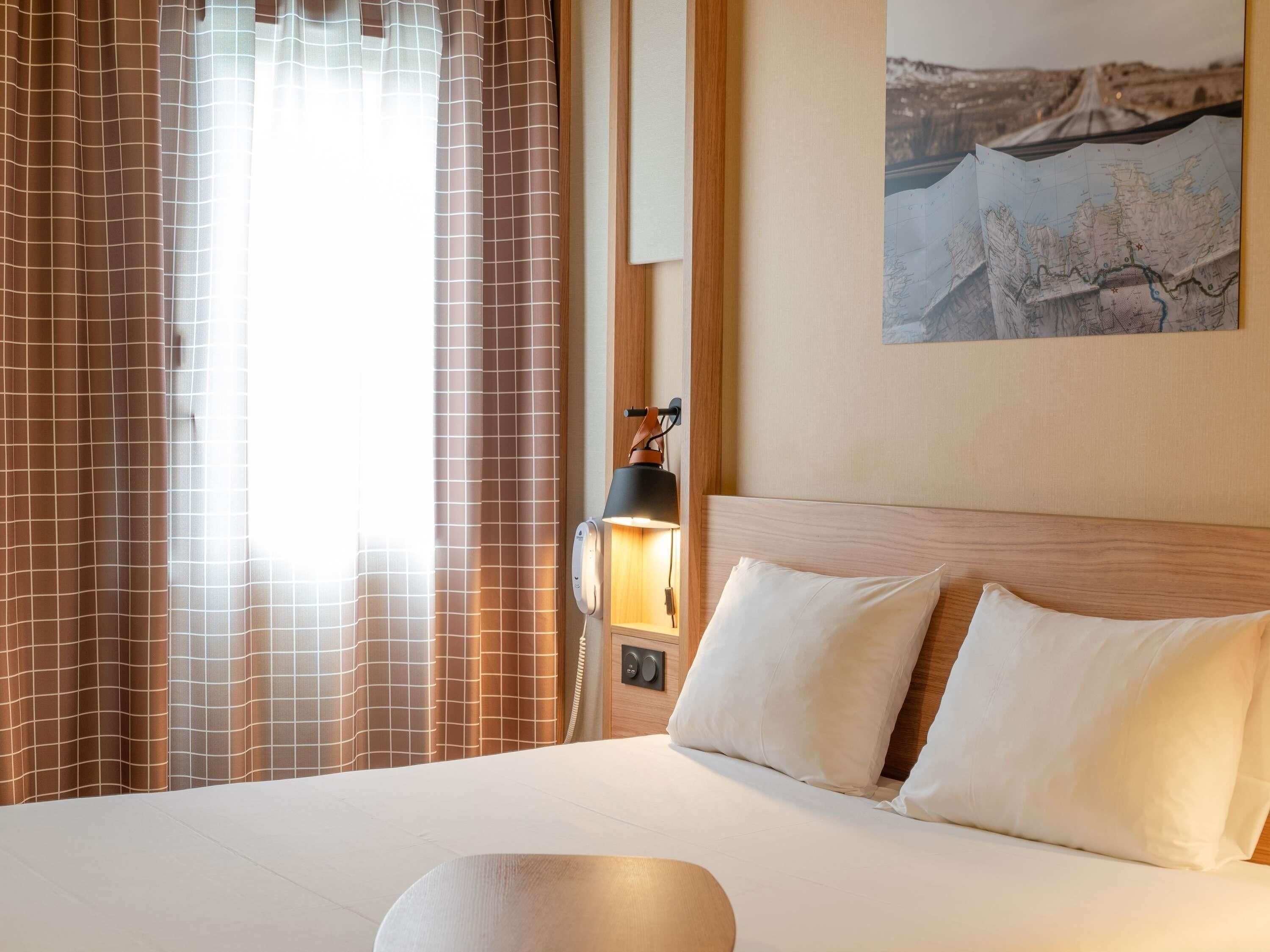 Et hotelværelse med seng, gardiner, lampe, vægmonteret telefon og et kort.