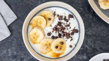 Uma tigela de iogurte com fatias de banana, pedaços de chocolate e bolinhas de chocolate branco sobre uma superfície texturizada.