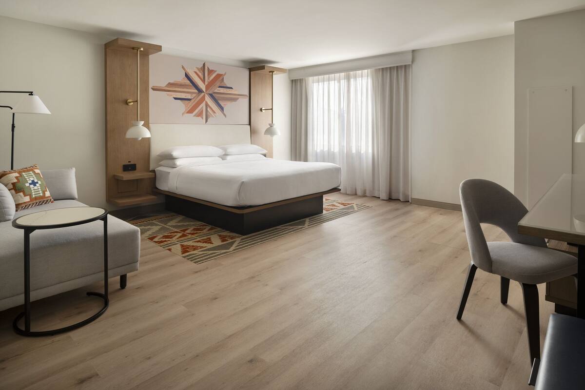 A modern hotel room with a large bed, a desk, a chair, and a small table.