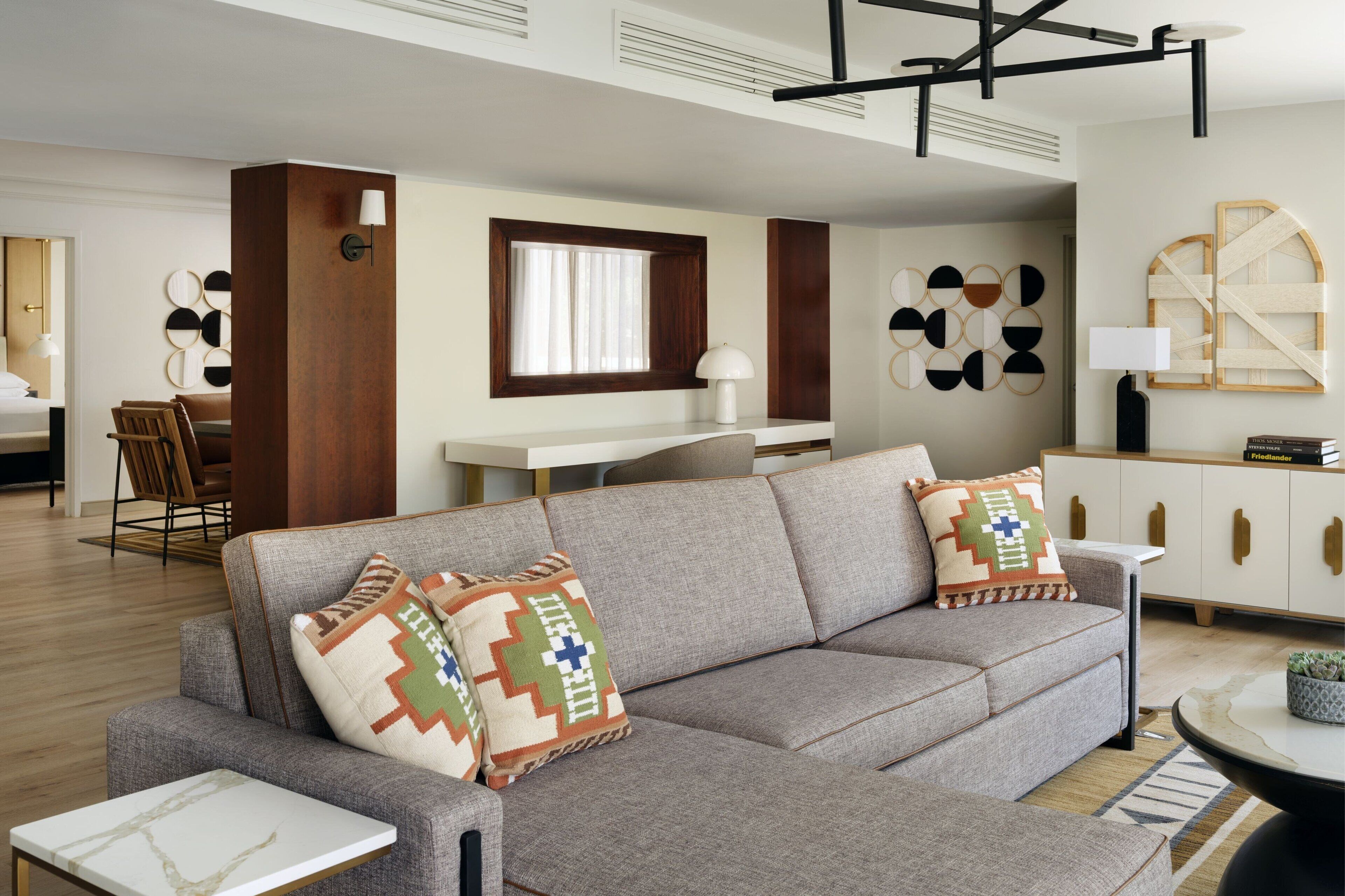 A modern living room with a grey sectional sofa, a small white table, and a round coffee table. There is a wooden cabinet with a mirror and a lamp. The room has a wooden floor and a large window with blinds.
