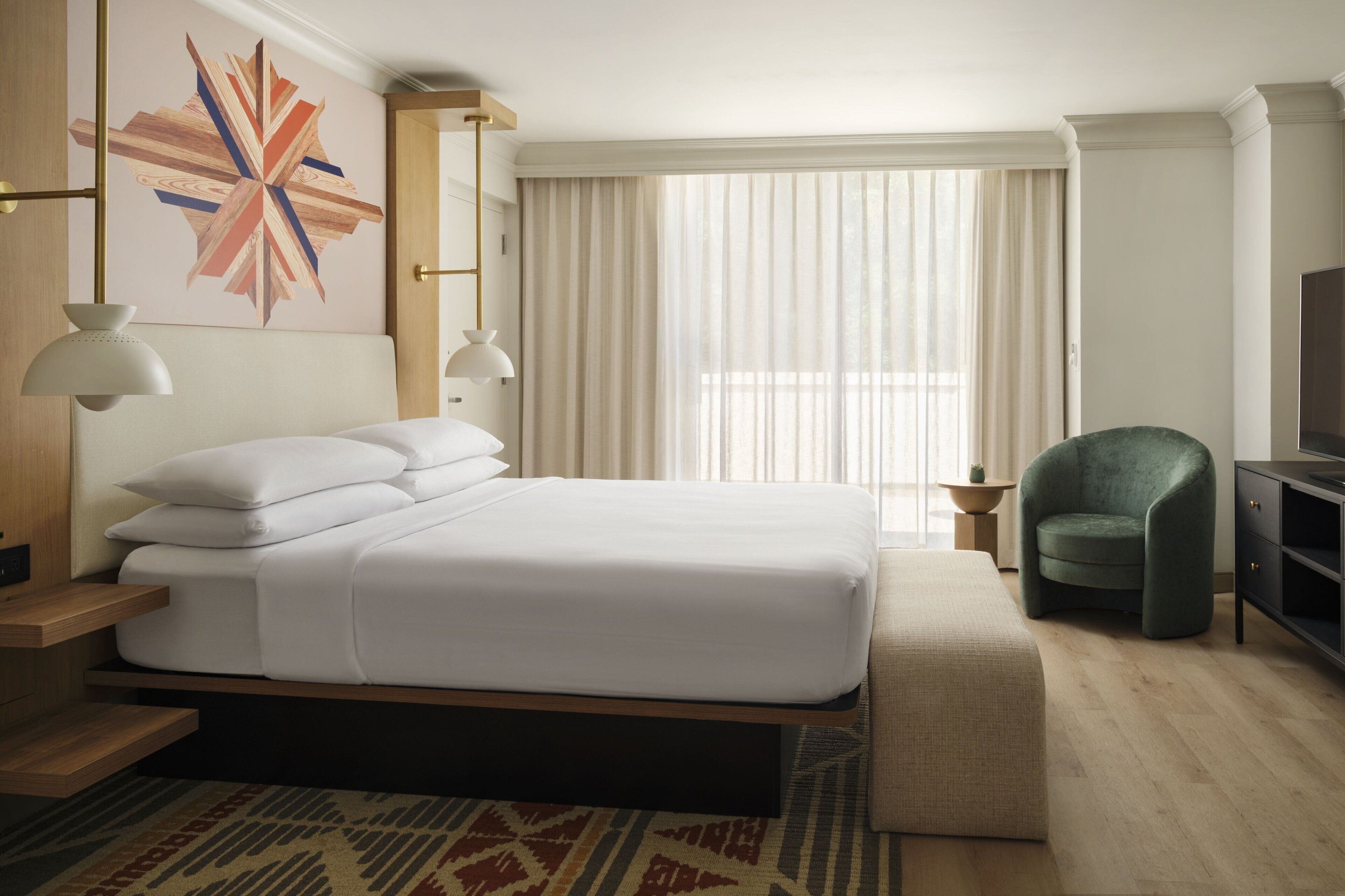 Habitación de hotel con una cama grande, lámparas de noche, un sillón verde, un televisor y una ventana con cortinas.