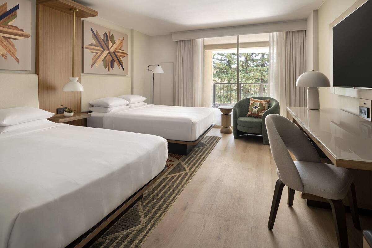 A hotel room with two beds, a desk, a chair, a TV, and a balcony with a view of trees.