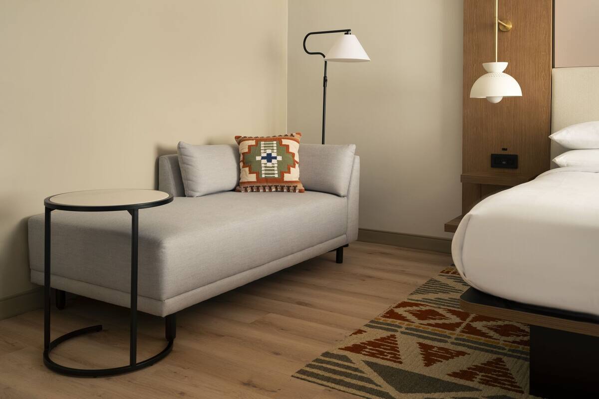 A modern hotel room with a grey sofa, a small round table, a patterned throw pillow, a floor lamp, and a bed with white bedding.
