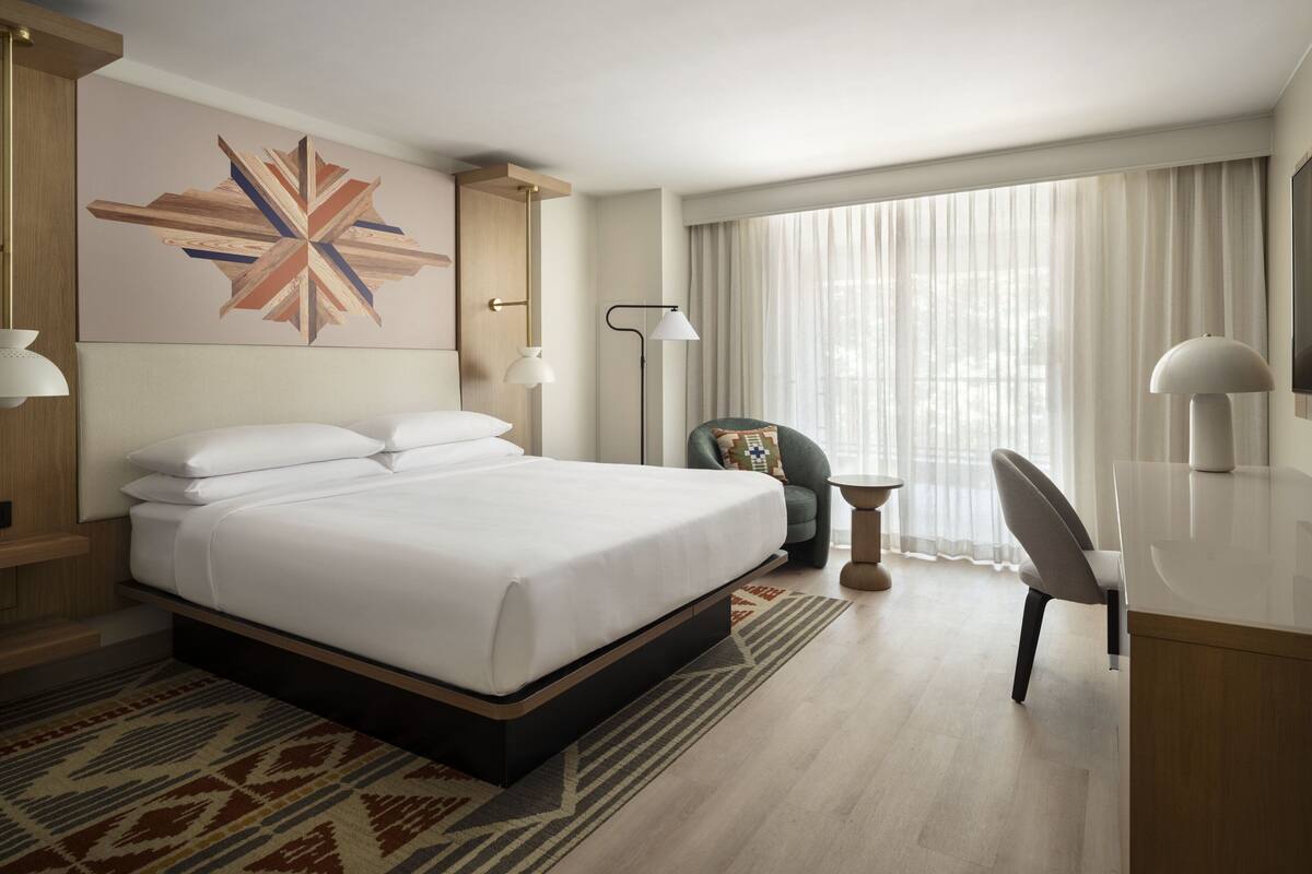 A hotel room with a large bed, a desk, a chair, a lamp, and a window with curtains.