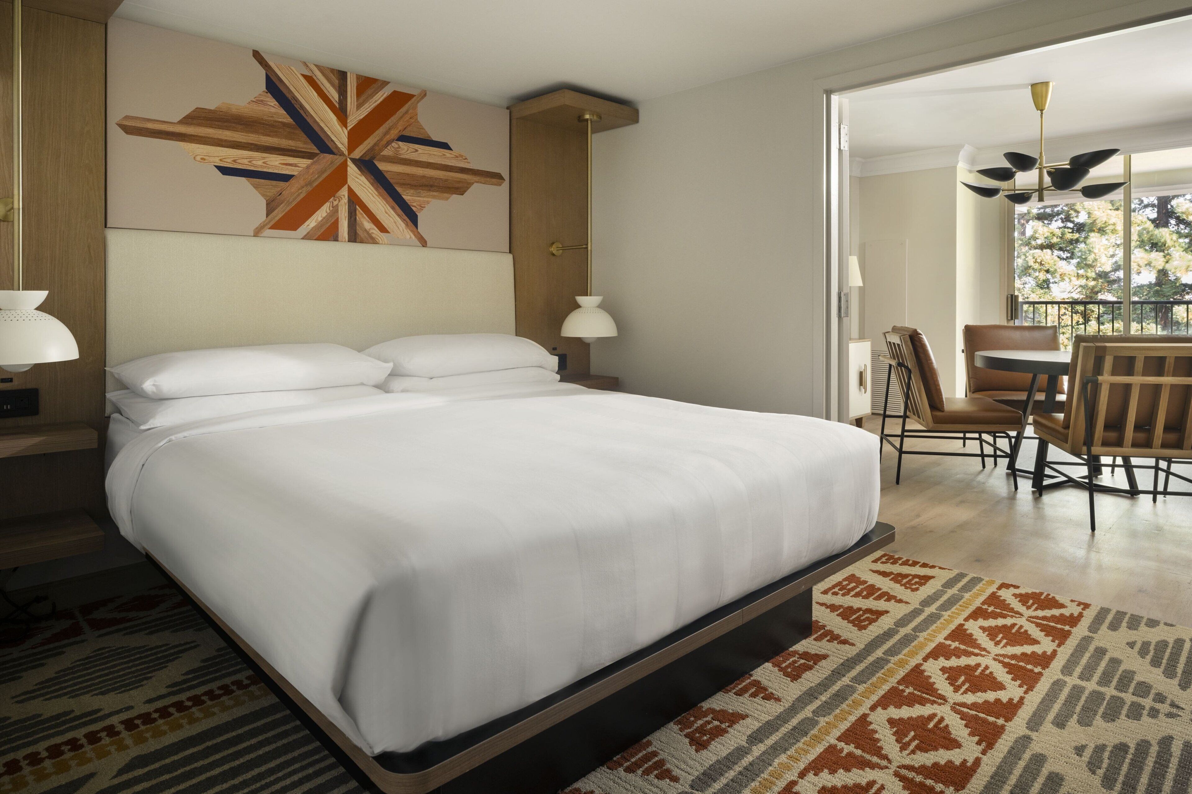 A neatly made bed with a patterned rug, a wall art piece, and a dining area with chairs and a table.