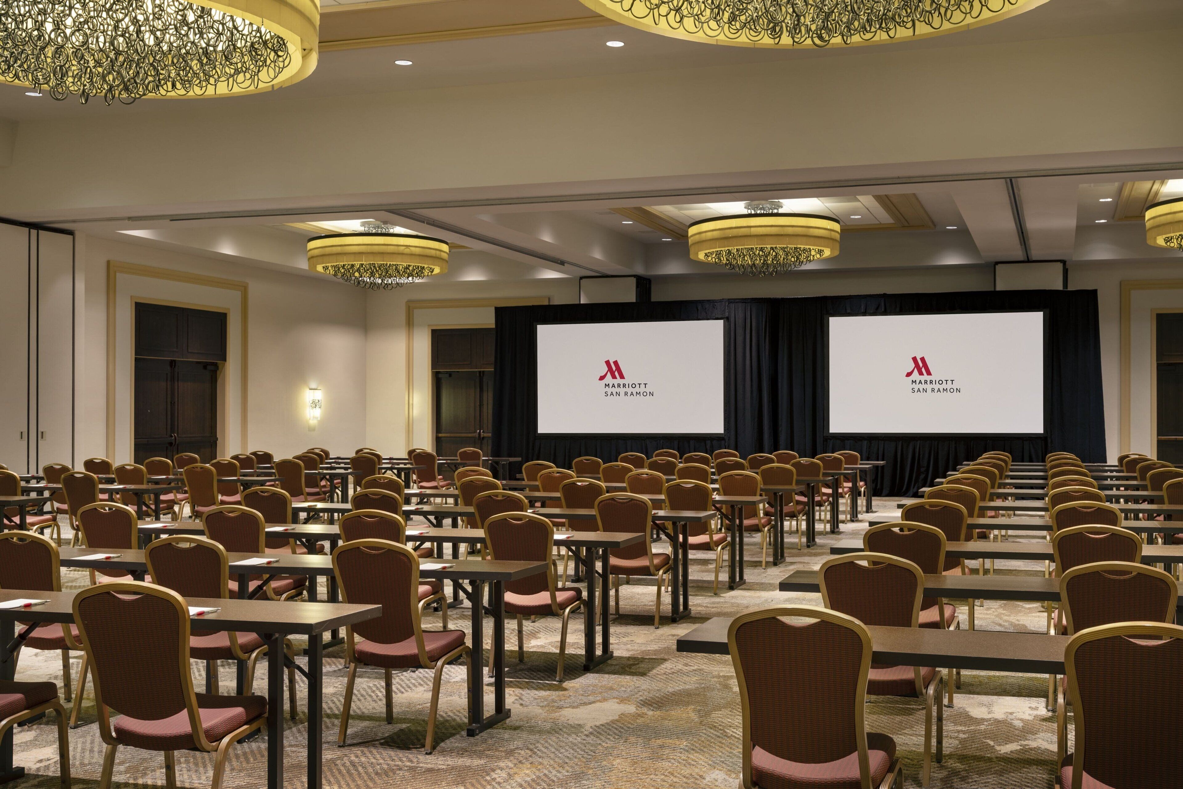 A conference room with rows of tables and chairs, a large screen, and a chandelier.