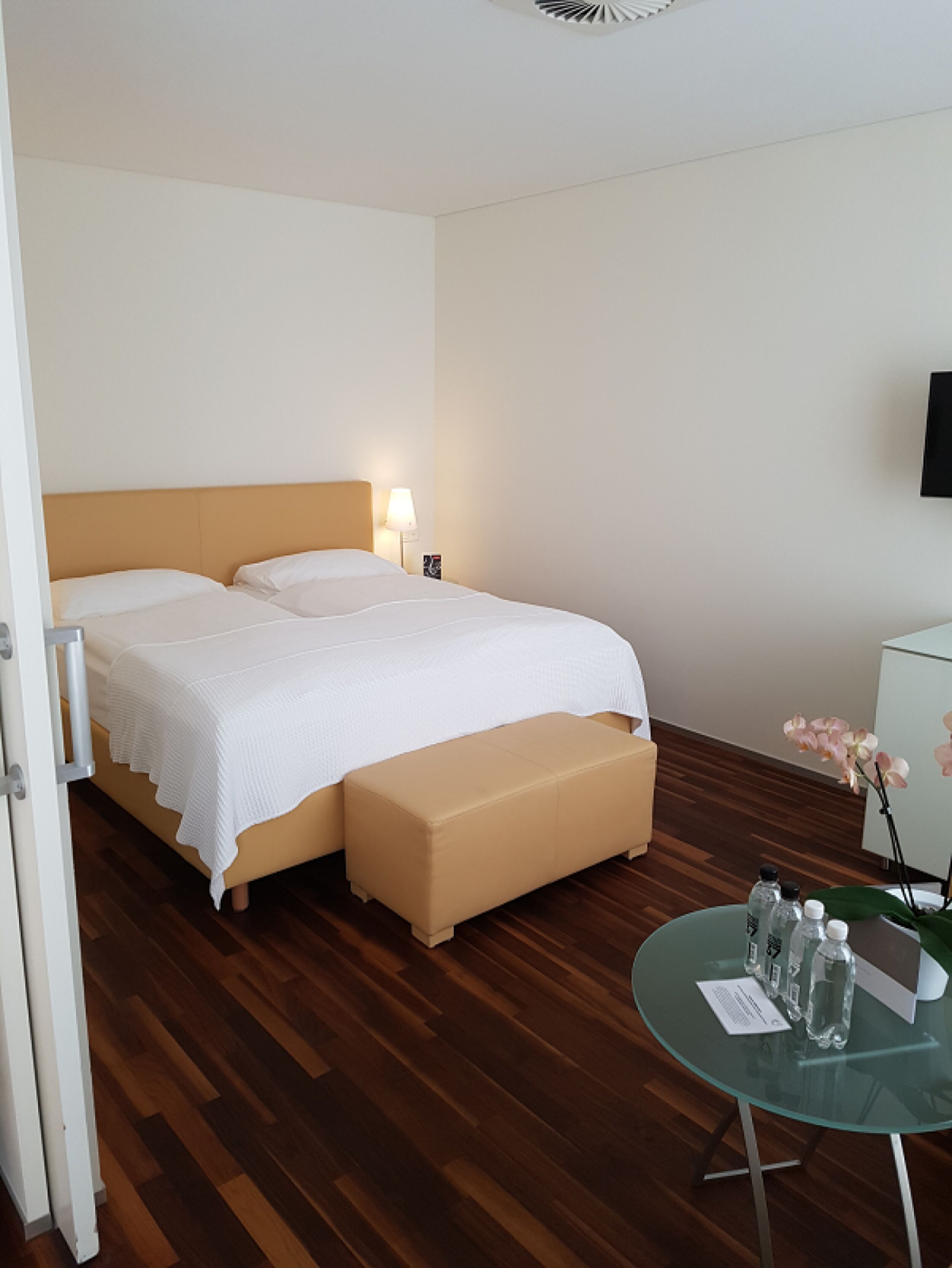 Una habitación de hotel con una cama, un banco, una mesa de vidrio con botellas y un mueble pequeño.