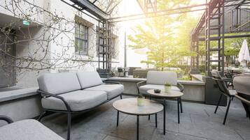 An outdoor seating area with a sofa, tables, and chairs, surrounded by a glass enclosure with a view of trees and a building.