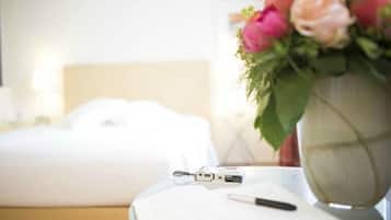 Una cama bien hecha con sábanas blancas, una mesita de noche con un bolígrafo, un pequeño dispositivo electrónico y un jarrón con flores rosadas.