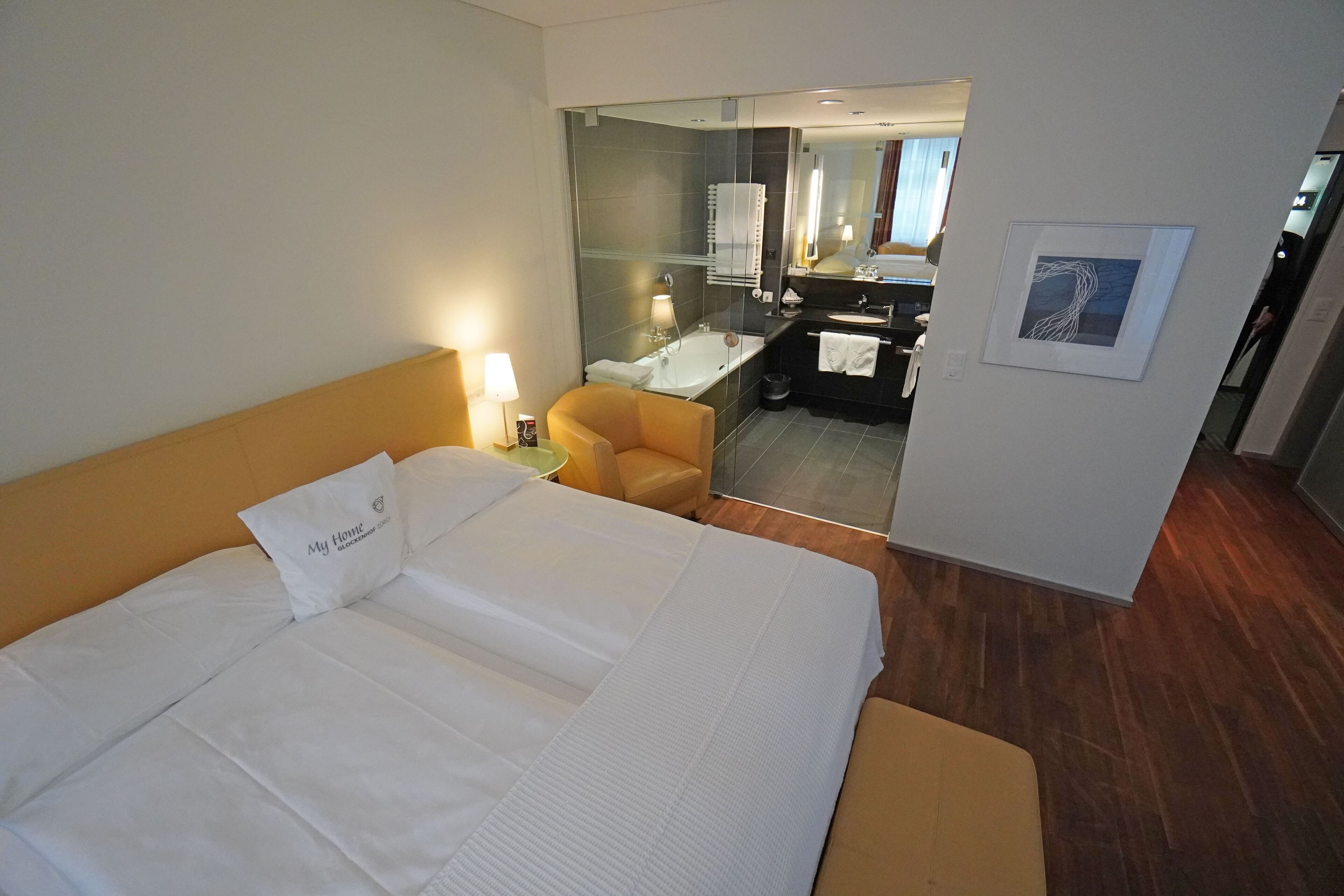 Camera d'albergo con un ampio letto, una poltrona, una scrivania e un bagno visibile tramite un pannello di vetro.