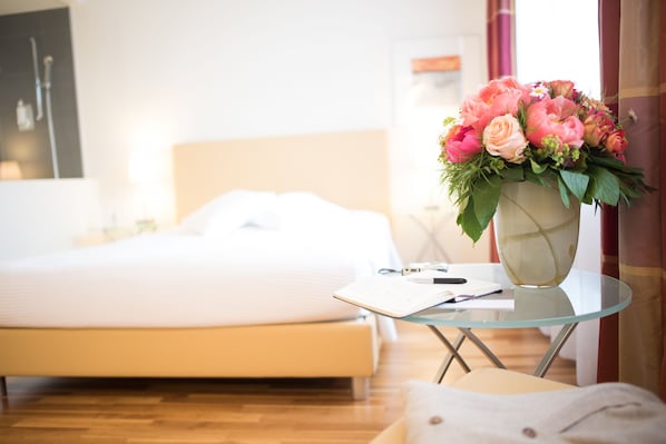 Una cama bien hecha con una colcha blanca, una mesa de vidrio con un florero y una mesita de noche con un cuaderno y un bolígrafo.