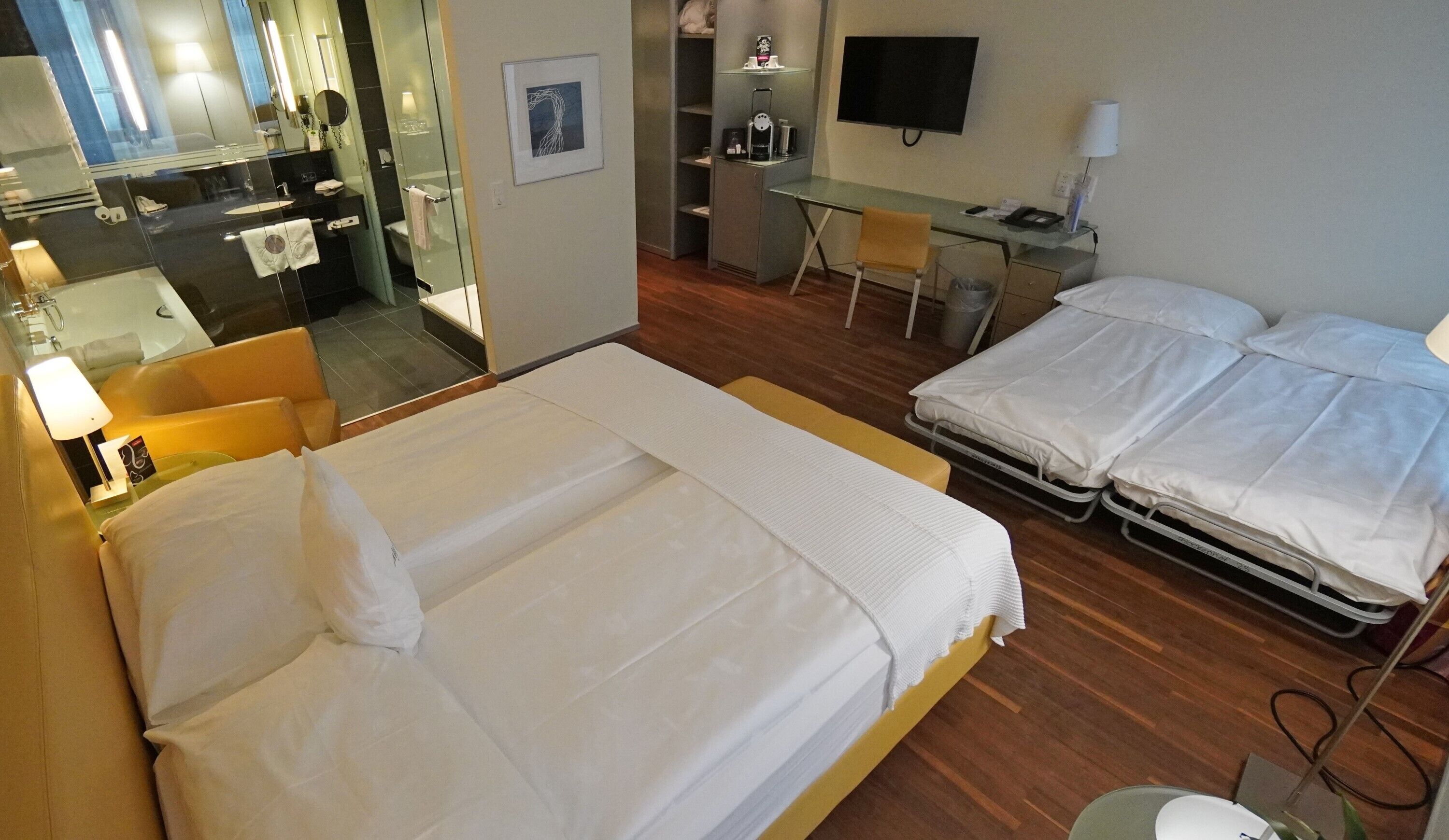Et hotelværelse med to senge, et skrivebord, en stol, et fjernsyn og et badeværelse, der kan ses gennem en glasvæg.