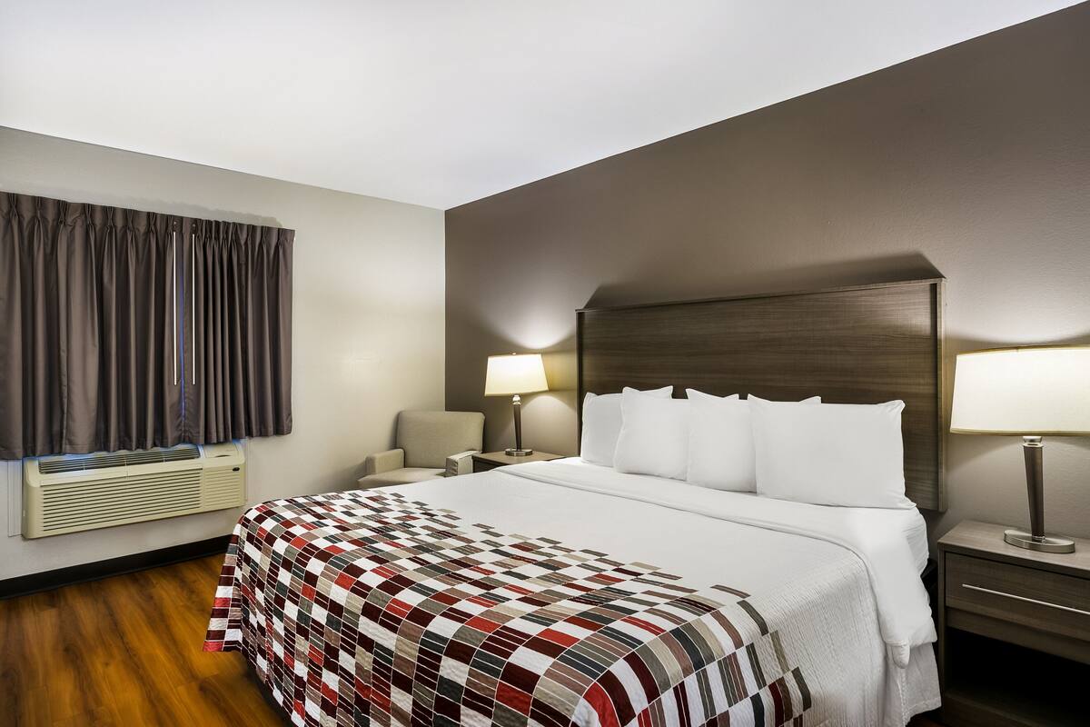 A hotel room with a large bed, two bedside tables with lamps, a chair, a window with curtains, and an air conditioning unit.