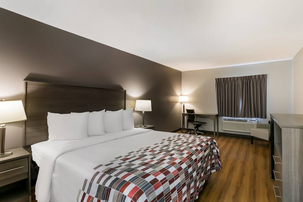 A hotel room with a large bed, two bedside tables with lamps, a desk with a chair, an air conditioning unit, and a TV on a cabinet.