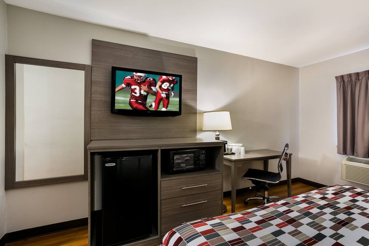 A hotel room with a flat-screen TV mounted on the wall, a desk with a chair, a microwave, a refrigerator, and a bed with a checkered bedspread.