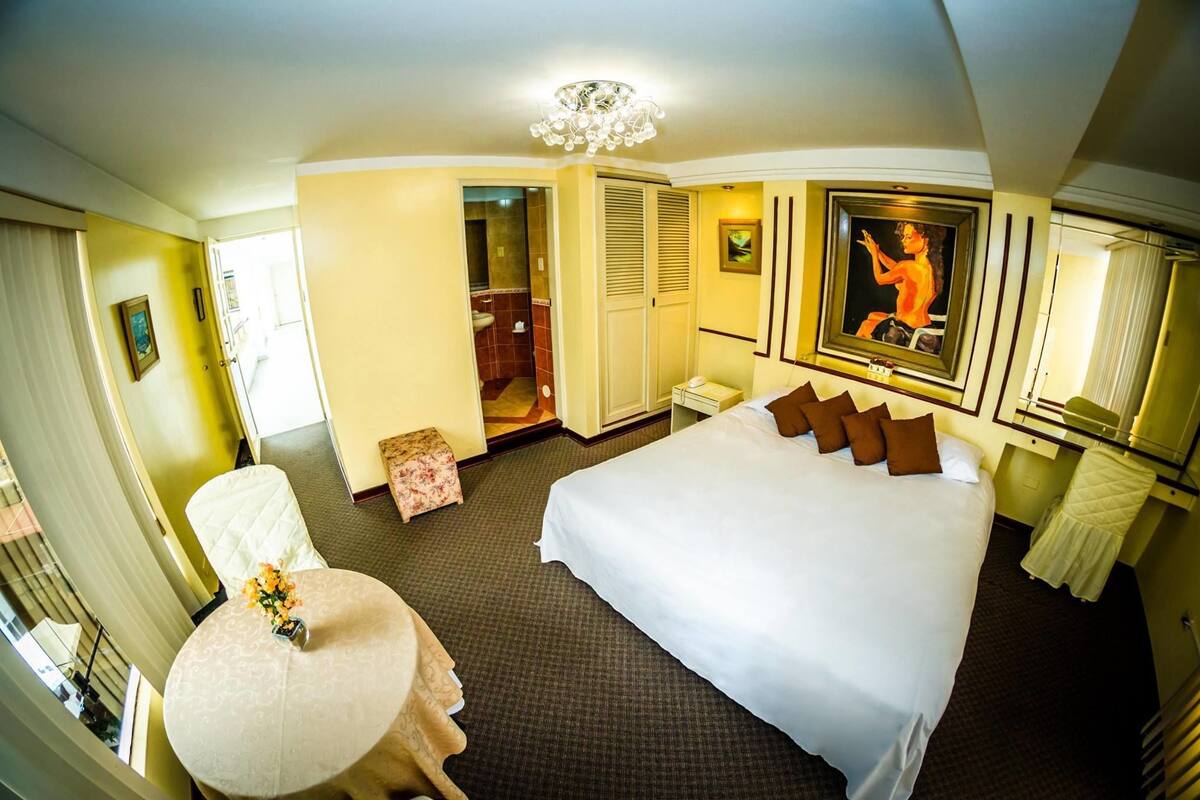 Habitación de hotel con una cama grande, una mesa redonda con un florero, una silla, un baño con ducha y un cuadro en la pared.