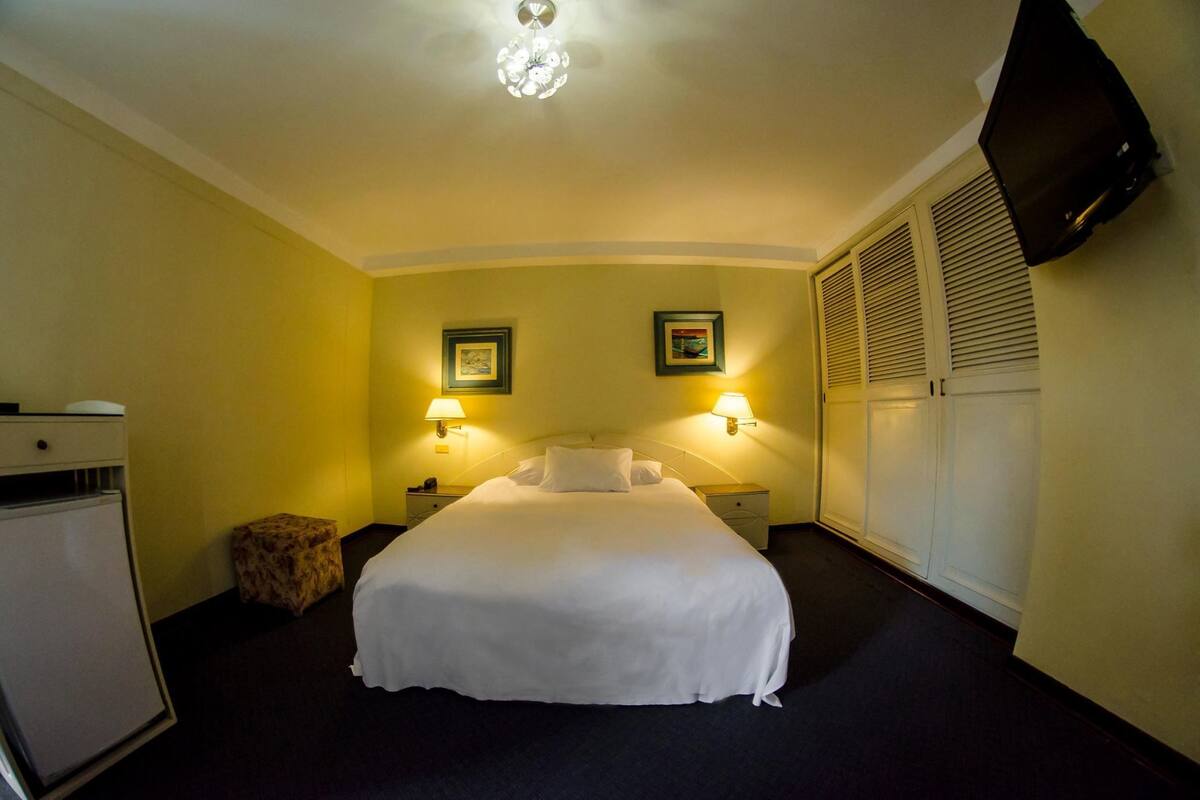 Una habitación de hotel con cama, lámparas de noche, televisión y un refrigerador pequeño.