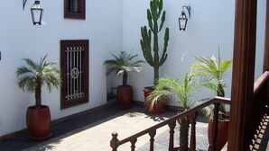 Une cour agrémentée de plantes en pot, un escalier en bois et une porte surmontée d’un portail en métal.