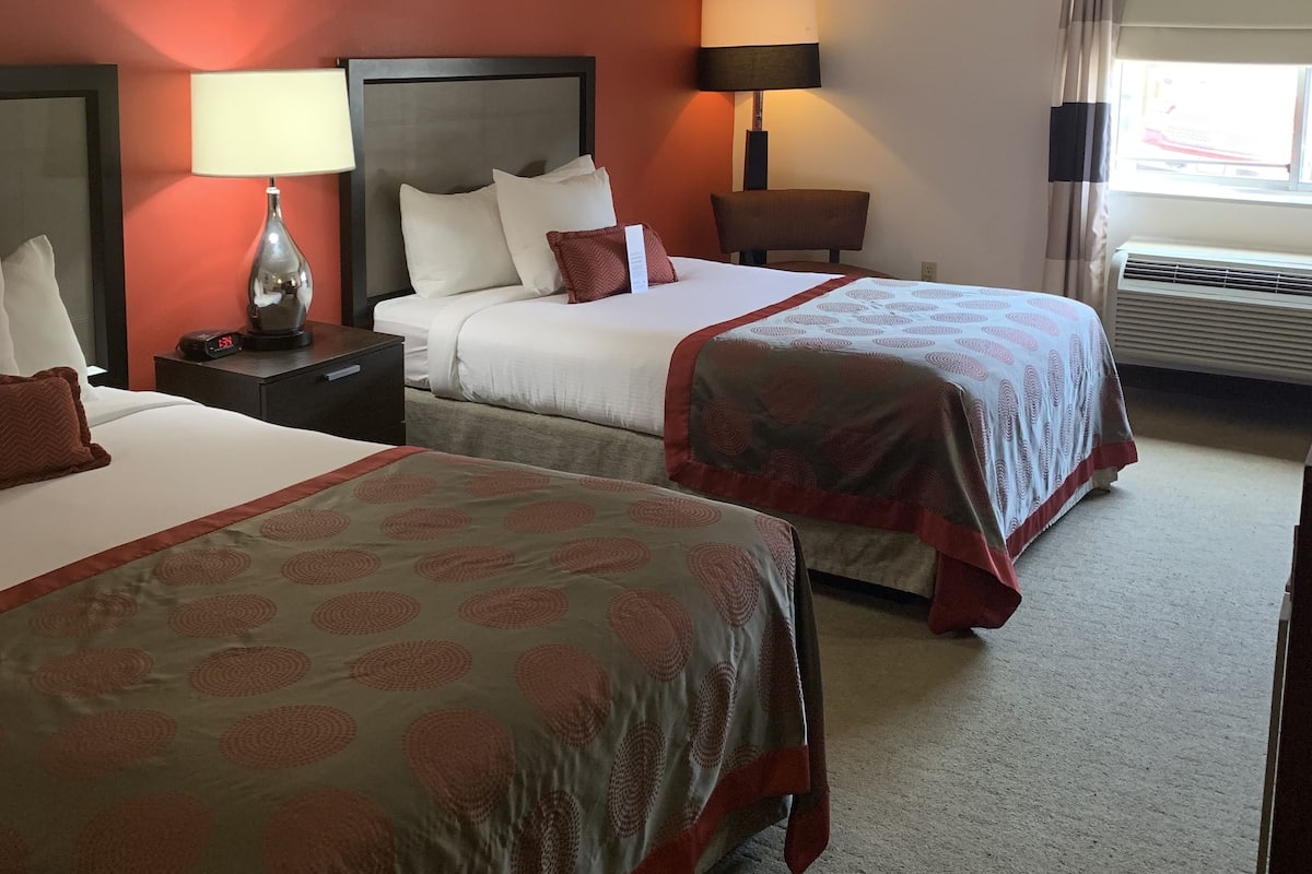 A hotel room with two beds, a nightstand with a lamp, a wall clock, and a window with curtains.